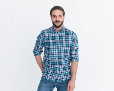 Ekose gömlekli seksi, tıraşsız bir adam ceplerinde el ele tutuşuyor ve gri arka planda havalı bir poz veriyor.