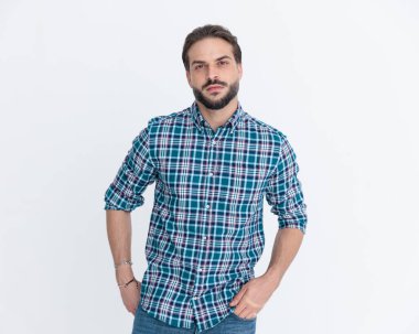 Çekici sakallı, kareli gömlekli, ceplerinde el ele tutuşan ve stüdyoda gri arka planda poz veren çekici bir adam.