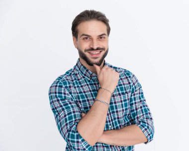 Ekose gömlekli mutlu genç adam stüdyoda gri arka planda gülümserken kollarını kavuşturuyor ve çenesine dokunuyor.