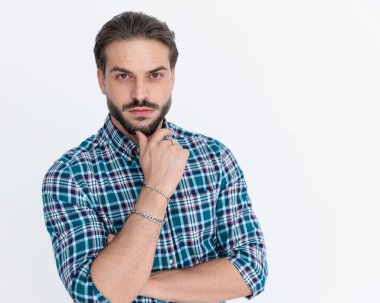 Ekose gömlekli düşünceli genç adam stüdyoda gri arka planda poz verirken çenesine dokunuyor ve düşünüyor.