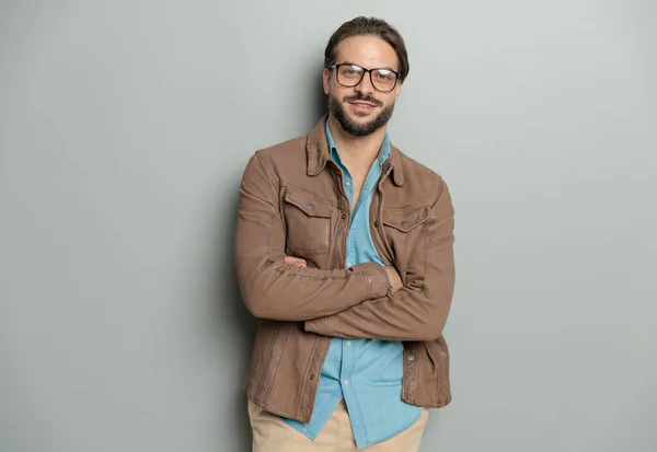 attractive bearded man with glasses wearing denim shirt and brown leather jacket crossing arms and smiling in front of grey background