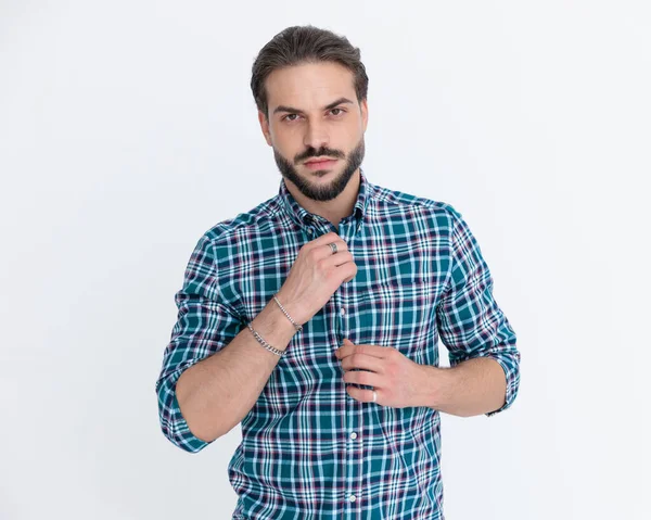 Seksi genç adam, sakallı, sıradan ekose gömleğini çözüyor. Sarı arka planda poz veriyor. 
