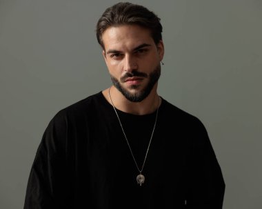 Siyah tişörtlü, sakallı, kendinden emin ve koyu gri arka planda poz veren seksi adam.