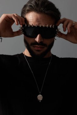 havalı genç adam güneş gözlüğü takıyor ve koyu gri arka planda poz verirken ileriye bakıyor.