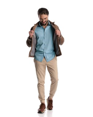 Gözlüklü yakışıklı genç adam beyaz arka planda yürürken kahverengi ceketini çekiyor ve düzenliyor. 