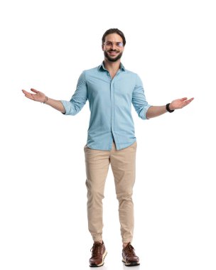 Gözlüklü gururlu genç adam kollarını açıyor ve gülümsüyor ve beyaz arka planın önünde duruyor.