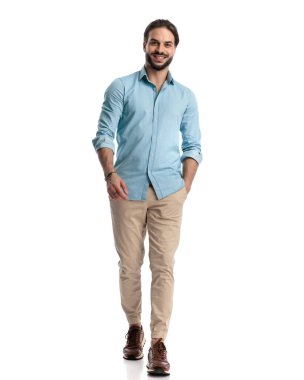 Sakallı yakışıklı genç adam elini cebine atıyor ve mutlu bir şekilde yürüyor ve beyaz arka planda gülümsüyor.