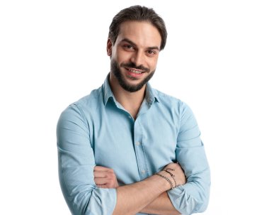 Sakallı, kot gömlekli, kollarını kavuşturup beyaz arka planda gülümseyen çekici bir genç. 