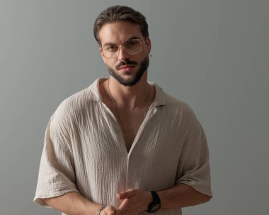 Gözlüklü, yakışıklı, tıraşsız bir adam. Stüdyoda gri arka planda kendine güvenen bir poz veriyor.