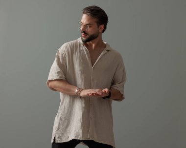 Gözlüklü, avuçlarını ovuşturan ve gri arka planın önünde duran seksi moda adamı. 