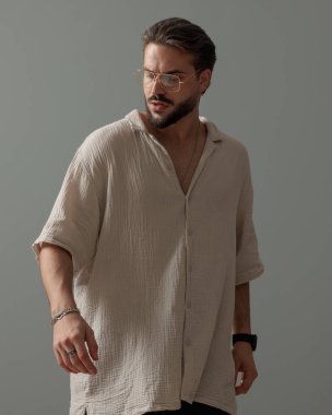 Gözlüklü seksi genç adam aşağı bakıyor ve gri arka planda poz veriyor.