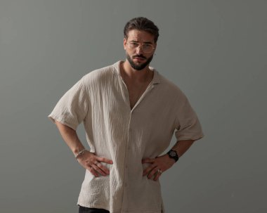 Gözlüklü yakışıklı adam, kalçasında elele tutuşuyor ve gri arka planda kendine güvenen bir şekilde poz verirken ileriye bakıyor.