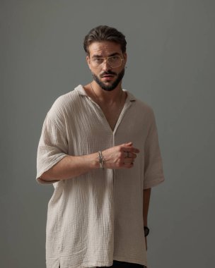 Gözlüklü yakışıklı sakallı adam gri arka planda poz veriyor.