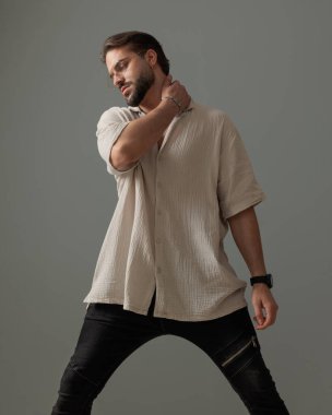 Tıraşsız, gözlüklü, boynunun arkasında el ele tutuşan ve gri arka planın önünde duran havalı bir adam. 