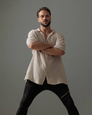 Yakışıklı, kendine güveni tam, gözlüklü, kollarını kavuşturup gri arka plana bakan bir adam.