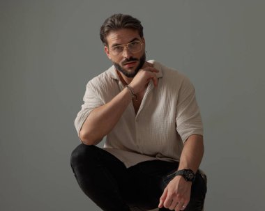 Gözlüklü çekici bir adam dizlerinin üstüne çömeliyor ve gri arka planda ileriye bakıyor. 