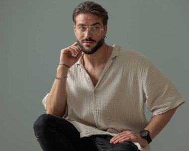 Dirseğini dizine dayayıp, gri arka planı düşünürken gözlerini kaçıran gözlüklü genç adam. 