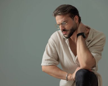 Havalı sakallı, gözlüklü, eli boynun arkasında poz veren ve diziyle gri arka planda poz veren havalı bir adam. 