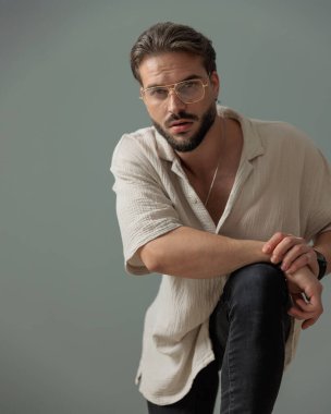 Gri arka planda diziyle poz verirken kollarını tutan ve ileriye bakan gözlüklü kendine güvenen genç adam.