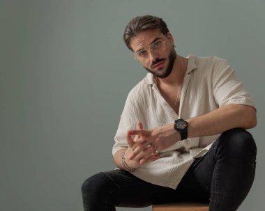 Gözlüklü çekici genç adam gri arka planda dirsekleriyle oturuyor ve poz veriyor.
