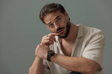 Gözlüklü, dizlerinin üstünde dirseklerini tutan ve gri arka planda ileriye bakan çekici bir adam. 