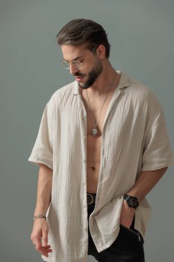 Gri arka planda el ele tutuşan ve aşağı bakan, gözlüklü, kendine güvenen sakallı adam.