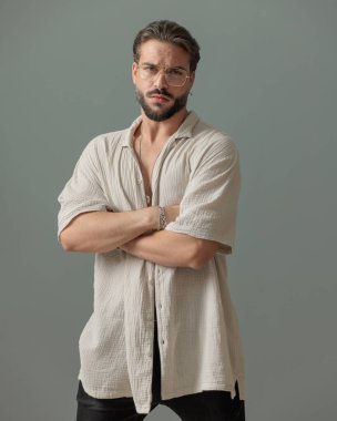 Gözlüklü, kollarını kavuşturmuş ve gri arkaplan önünde poz veren seksi genç adam.