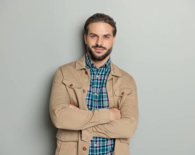 Bej ceketli, kollarını kavuşturup gri arka planda gülümseyen mutlu bir adamın portresi.