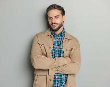 Ceketli yakışıklı, sakallı, sıradan bir adam kollarını kavuşturuyor ve gri arka planda ileriye bakıyor.