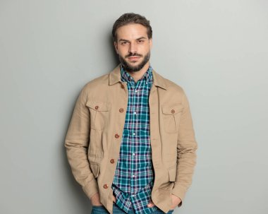 Bej ceketli, cebinde ekose gömleği olan ve gri arka planda poz veren yakışıklı sıradan bir adamın portresi.