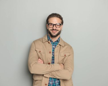 Kollarını kavuşturup gri arka planda poz verirken gülümseyen gözlüklü yakışıklı adam.