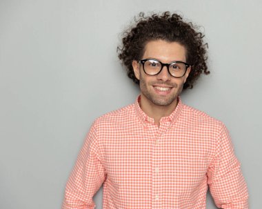 Ekose gömlekli, gülümseyen, mutlu, ayakta duran ve stüdyoda gri arka planda poz veren kıvırcık saçlı adamın portresi.