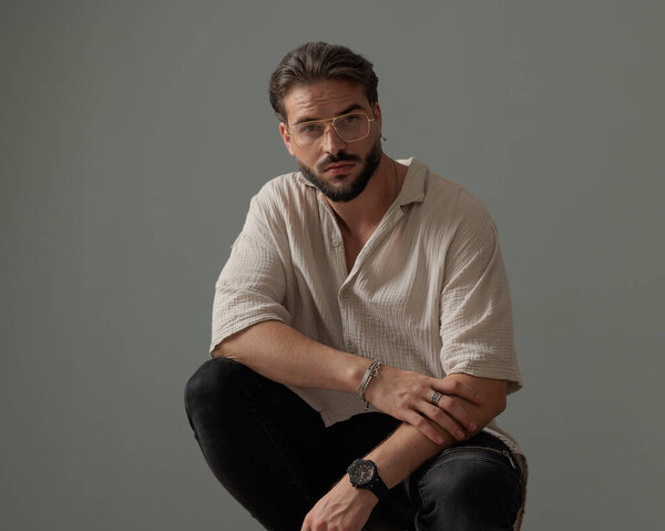 крутой модный парень в очках, держащий локти на коленях и скрежещущий перед серым фоном 