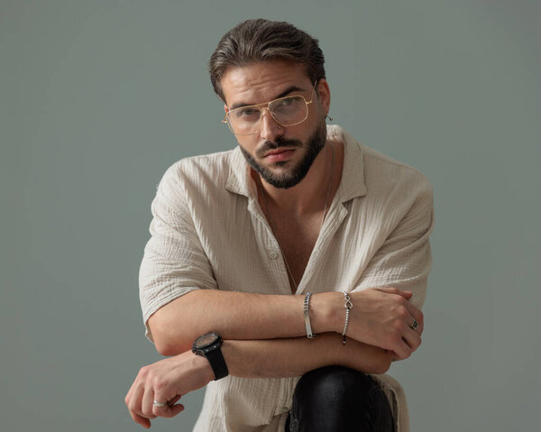 небритый молодой человек в очках скрещивая руки и держа колено перед серым фоном 