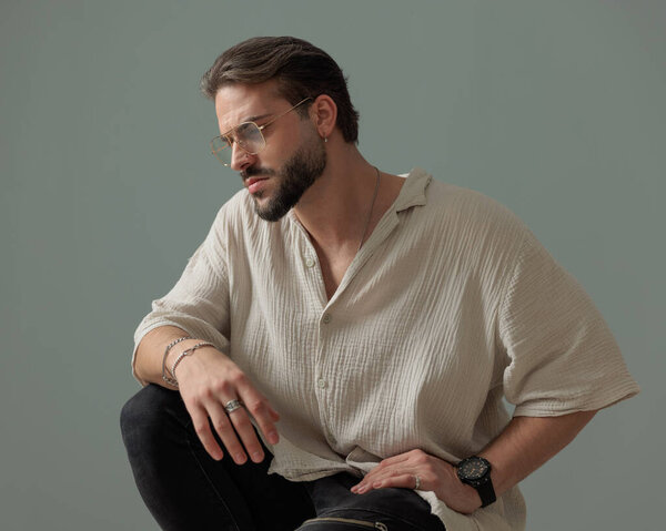 sexy unshaved man with glasses crouching with elbow on knee and looking to side in front of grey background 
