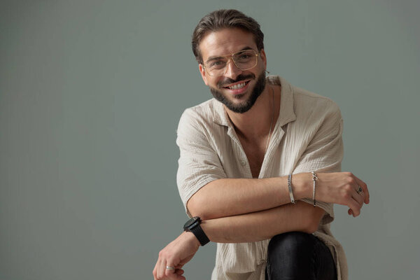 счастливый молодой человек в очках, держа колено и складывая руки, улыбаясь и глядя вперед на сером фоне