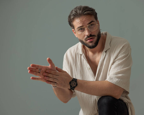 красивый парень в очках, держащий локоть на коленях и трогательные ладони, позируя на сером фоне 