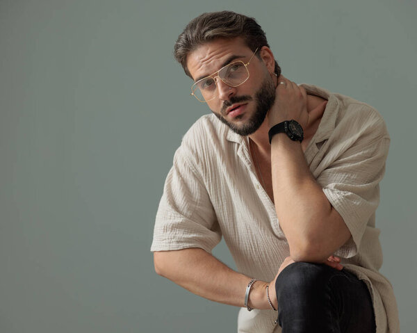 sexy bearded man with glasses holding hand behind neck and posing with knee up in front of grey background 