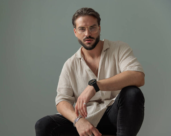 уверенный в себе небритый мужчина в очках сидит и позирует с локтями на коленях на сером фоне 