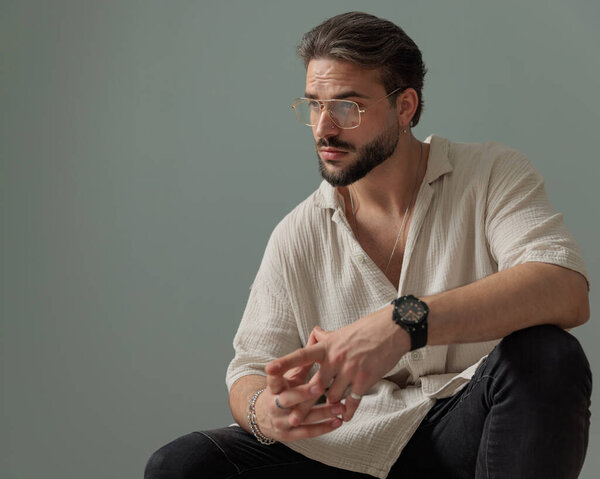 sexy young man with glasses holding elbows on knees and looking away in front of grey background 