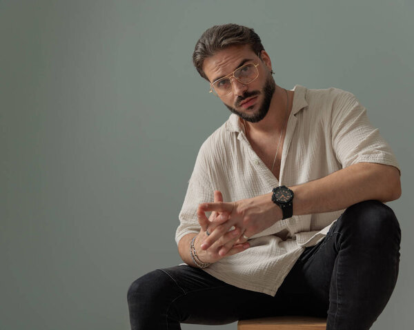 привлекательный молодой человек в очках сидит и позирует с локтями на коленях на сером фоне