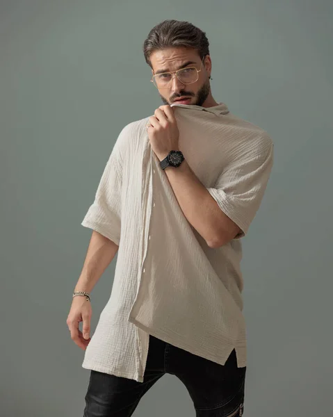 グラス付きセクシーな若い男は グレーの背景の前にポーズしながらシャツで顔を引っ張ってカバー — ストック写真