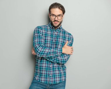 Kollarını kavuşturup gri arka planda poz veren gözlüklü havalı sıradan bir adam.