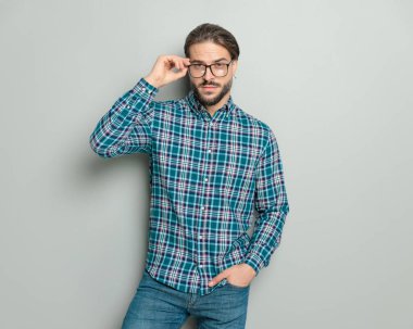 Ekose gömlekli sıradan bir genç adamın portresi. El ele tutuşup, gri arka planda durup poz verirken gözlükleri ayarlıyor.