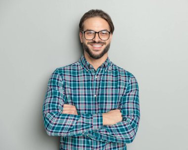 Gözlüklü gururlu bir gencin portresi kollarını kavuşturup stüdyoda gri arka planda poz verirken gülümsüyor.