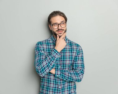 Kareli gömlekli, gözlüklü, kollarını kavuşturup gri arka planda düşünen dalgın bir adamın portresi.