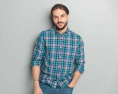 Kareli gömlek giyen, el ele tutuşan, gri arka planda poz veren kendinden emin bir adam.