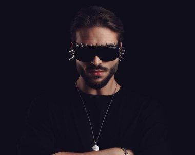 Tıraşsız, gözlüklü genç adam kollarını kavuşturup siyah arka planda ileriye bakıyor.