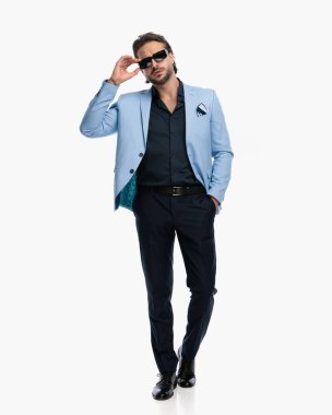 Tıraşsız, takım elbiseli, cebinde güneş gözlüklerini ayarlayan ve beyaz arka planda hareket eden zarif bir adam. 