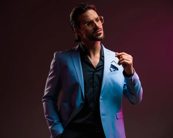 stock image portrait of elegant man with open collar shirt looking away and holding hand in pocket in front of black background with pink lights 